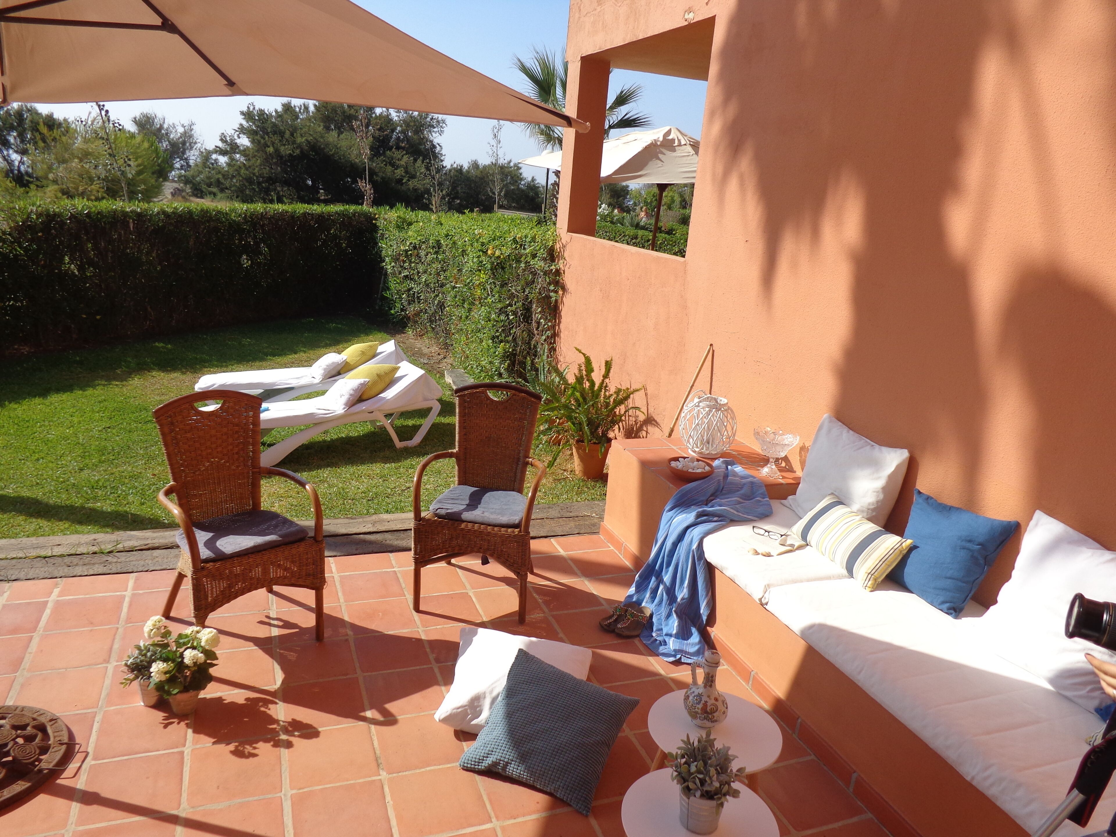 Eine sonnendurchflutete Terrasse mit einer Couch, zwei Korbstühlen, einem kleinen Tisch und einer Liege.