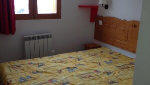 Une chambre à coucher avec un lit, une fenêtre, un radiateur, une étagère rouge et une lampe fixée au mur.