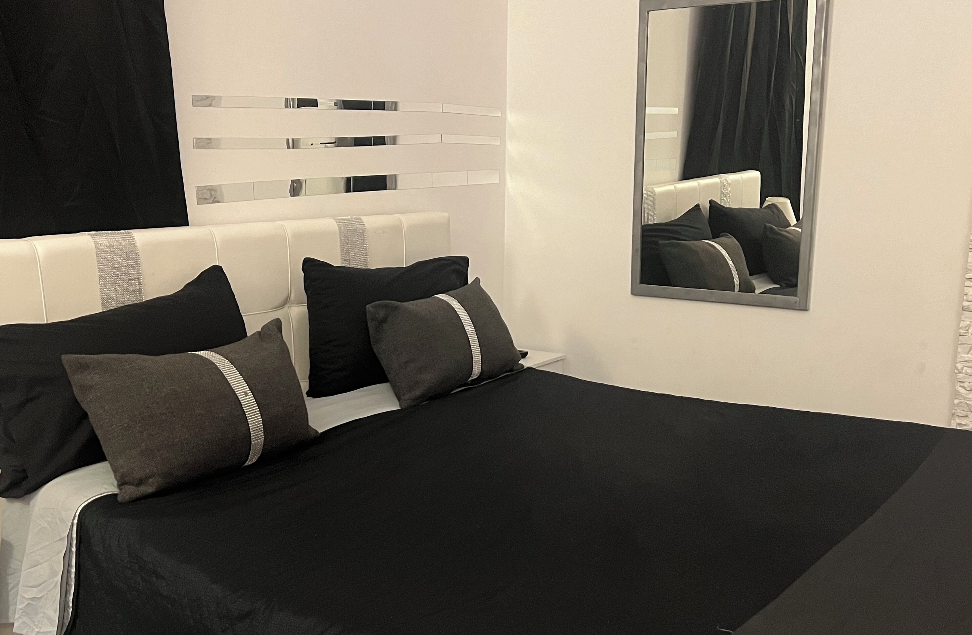 A modern bedroom with a white headboard, black bedding, and a mirror reflecting the room.