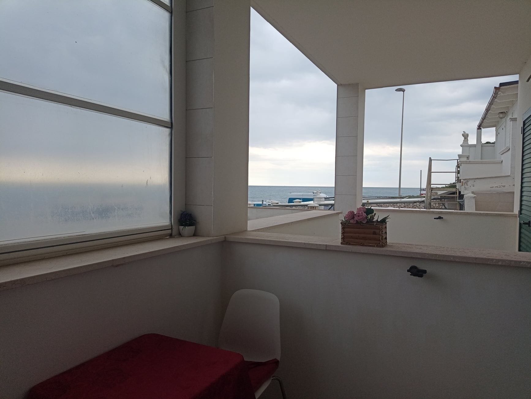 A balcony with a view of the sea, a red table, a white chair, and a small wooden box with flowers.