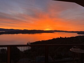 Ein Sonnenuntergang über einem stillen See mit Bergen im Hintergrund, Blick vom Balkon mit Geländer und Tisch.