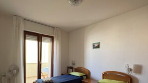 Una habitación con dos camas individuales, un ventilador de techo, una ventana con cortinas y una pequeña mesa con una silla.