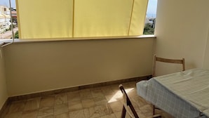 Un pequeño balcón con una toldilla amarilla, una silla de madera y una mesa con mantel a cuadros.