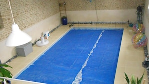 Une grande piscine intérieure au sol bleu, entourée d’une clôture blanche. On y trouve des plantes en pot et une petite échelle près de la piscine.
