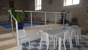 Un espace piscine intérieure doté d’un sol à carreaux, de chaises blanches et d’une table dressée pour les repas.