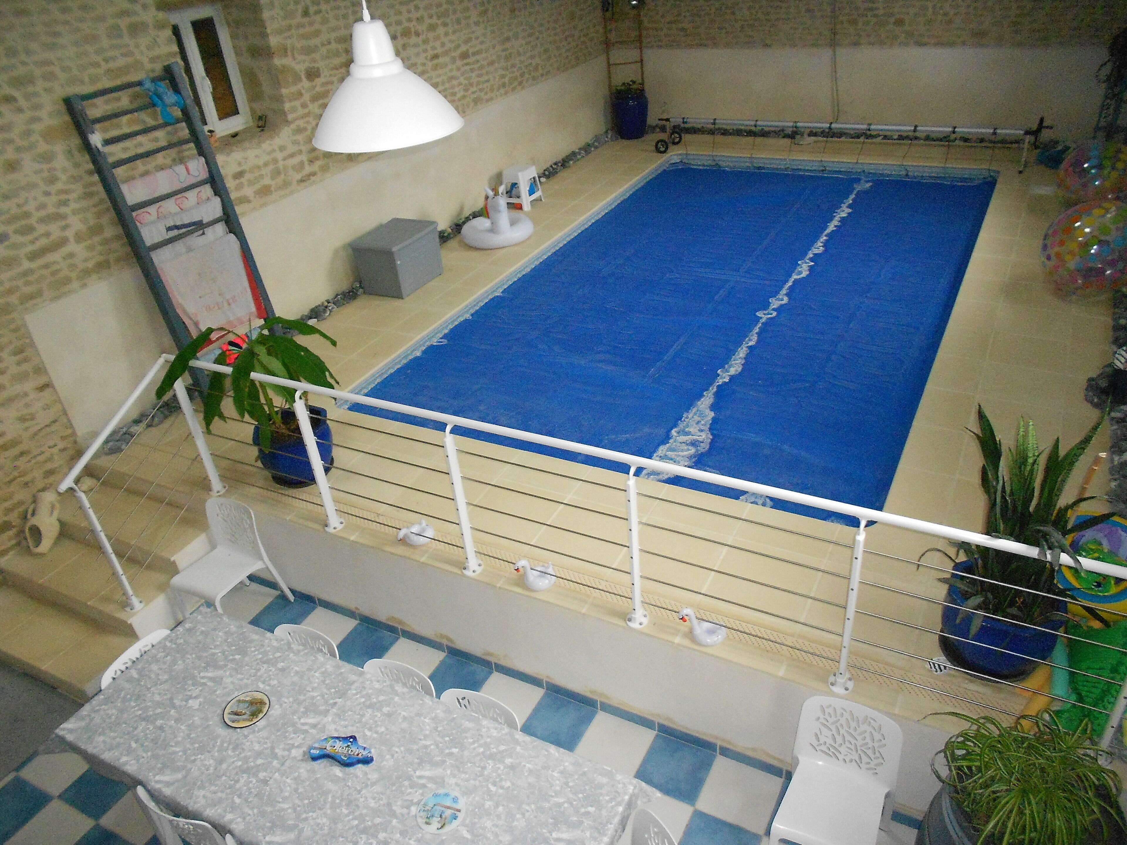 An indoor swimming pool with a blue water surface, surrounded by a white railing. There are potted plants and a small table with chairs in the foreground.