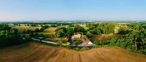 An aerial view of a rural landscape with a large estate featuring multiple buildings, including a prominent house with a red roof, surrounded by fields and trees.