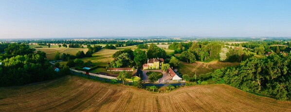 An aerial view of a rural landscape with a large estate featuring multiple buildings, including a prominent house with a red roof, surrounded by fields and trees.