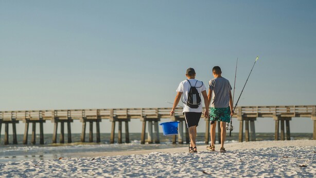 Dos personas caminando sobre una playa arenosa con un muelle al fondo.