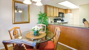 Un comedor con una mesa de vidrio, sillas de madera y un espejo en la parte superior. Se ve una cocina con armarios de madera y electrodomésticos al fondo.