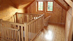 A wooden cabin with a high ceiling, a wooden crib, and two windows with a view of greenery.