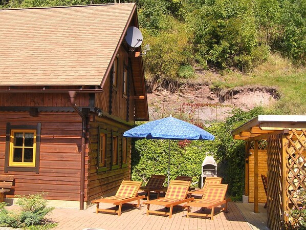 A wooden cabin with a satellite dish, a patio with wooden chairs, and a blue umbrella.