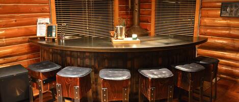 Interior de uma cabana de madeira com um espaço de bar central, incluindo uma mesa redonda, banquetas e uma pia.