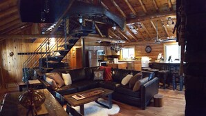 A rustic cabin interior with a wooden staircase, a living room with leather sofas, a dining area, and a kitchen.