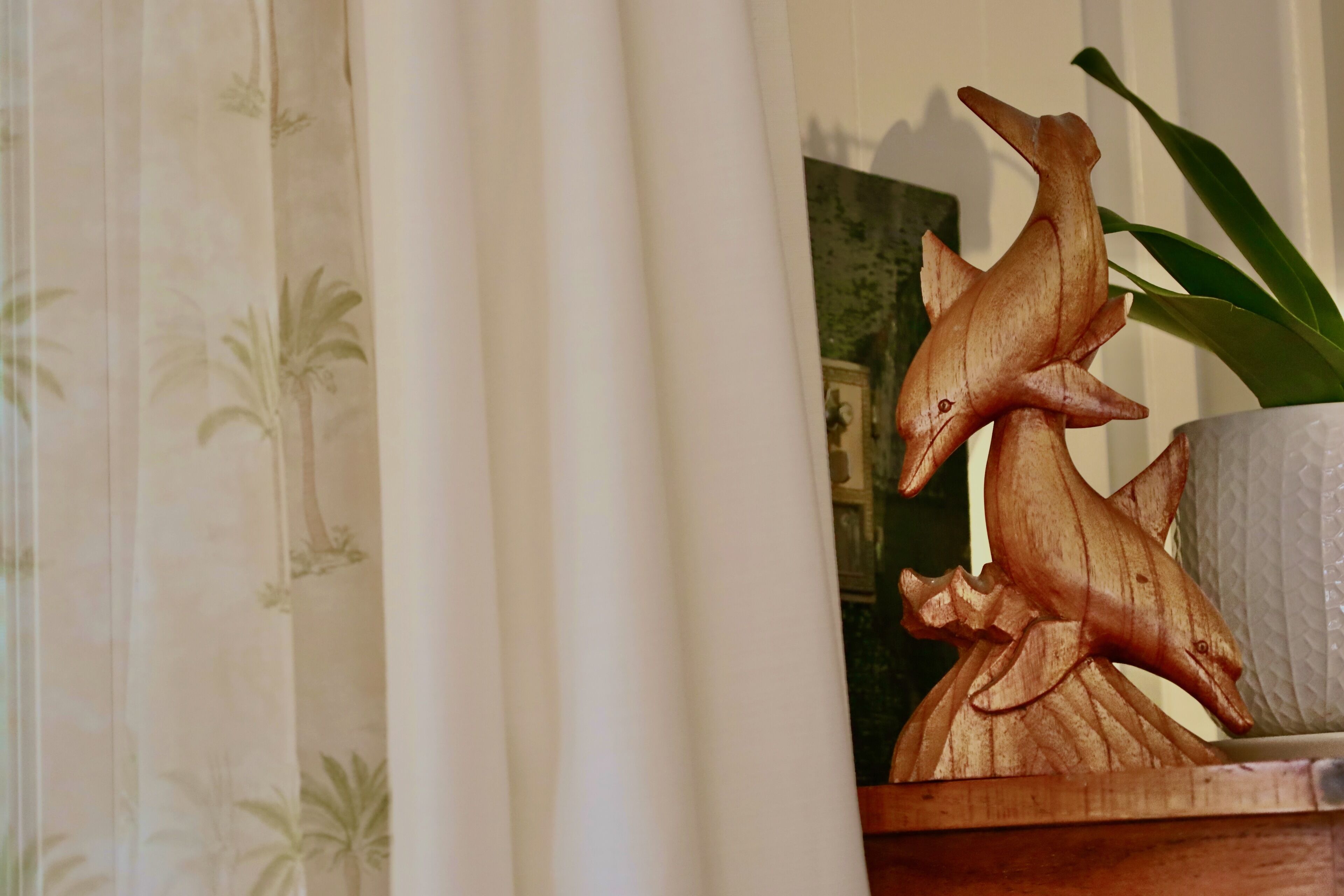 A wooden sculpture of a dolphin on a shelf, a potted plant, and a curtain with a palm tree pattern.