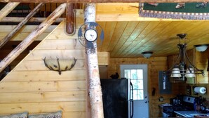 Interior de una cabaña de madera con una escalera, zona de comedor, cocina y sala de estar con sofá y cojines estampados.