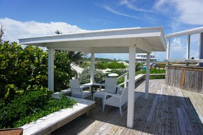 Un área de estar al aire libre con techo, con sillas y mesas blancas, con vista a una playa y el océano.