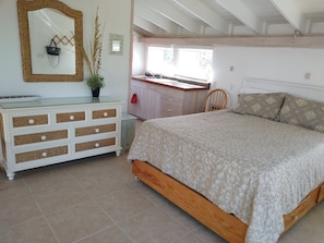 Une chambre avec un lit en bois, une commode blanche aux tiroirs en osier, un miroir et une fenêtre donnant sur l’extérieur.