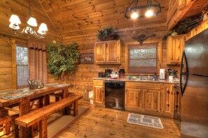 A wooden cabin interior with a dining table, benches, a kitchen with wooden cabinets, a refrigerator, and a chandelier. - Peaceful Woods #25- Charming & Clean Log Cabin! (Sevierville)