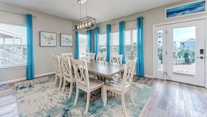 A dining area with a wooden table and chairs, a chandelier, and a view of the outside through large windows with blue curtains. - Steps From The Oceanfront! Pool, Pet Friendly, Wifi, king beds! WEEKLY DISCOUNTS (Cape San Blas)