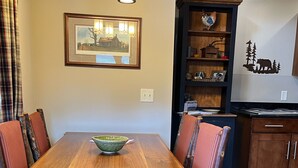 Une table à manger en bois avec des chaises, un lustre, un tableau encadré, un buffet en bois et un coin cuisine.