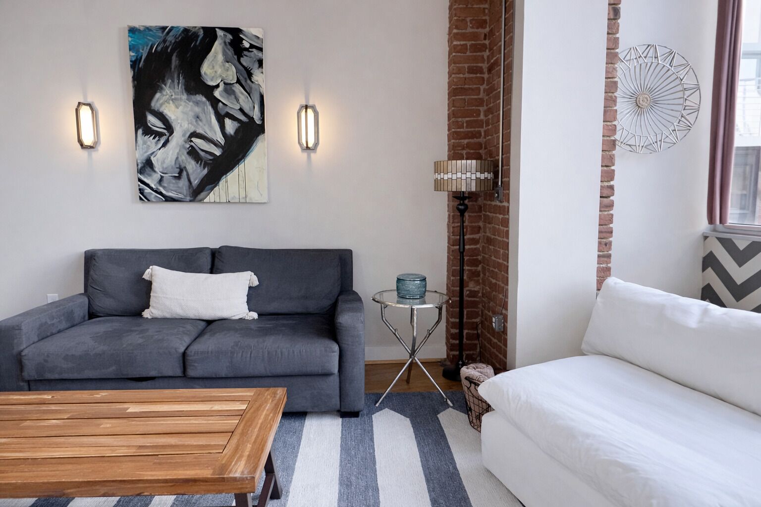 Una sala de estar moderna con un sofá gris, un sofá blanco, una mesa de centro de madera y un tapete a rayas. En la pared hay un cuadro, una lámpara de pie y un elemento decorativo.