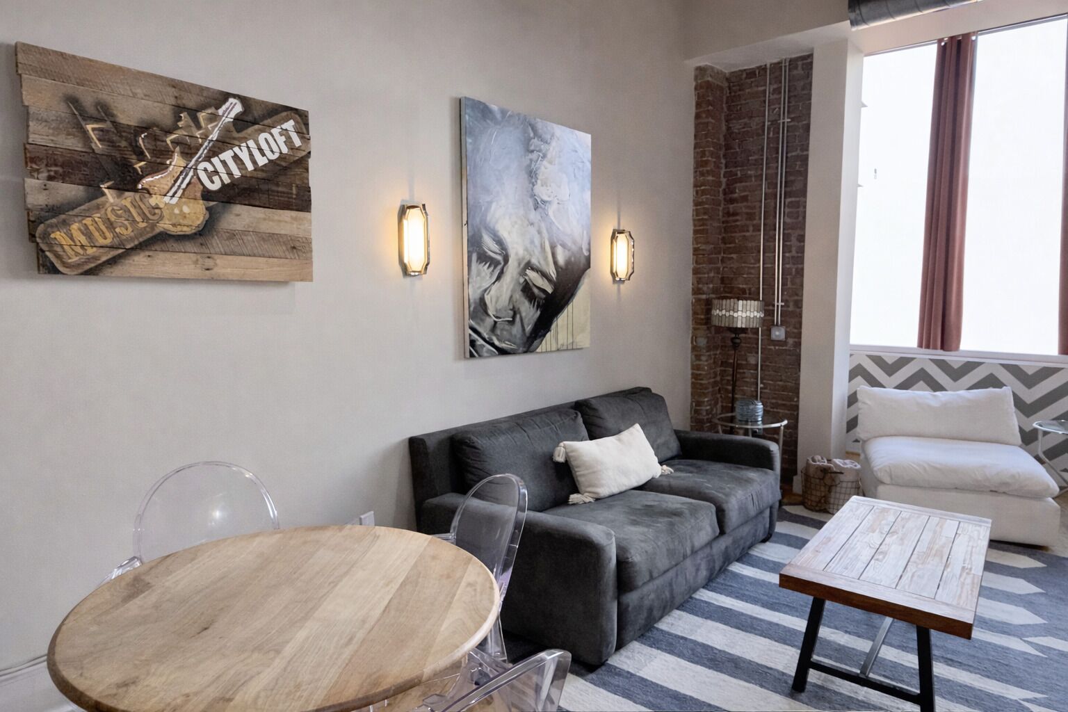 Una sala de estar moderna con un sofá gris, una mesa de centro de madera y una mesa de comedor redonda. Hay dos lámparas fijadas a la pared y un cuadro abstracto grande sobre el sofá.