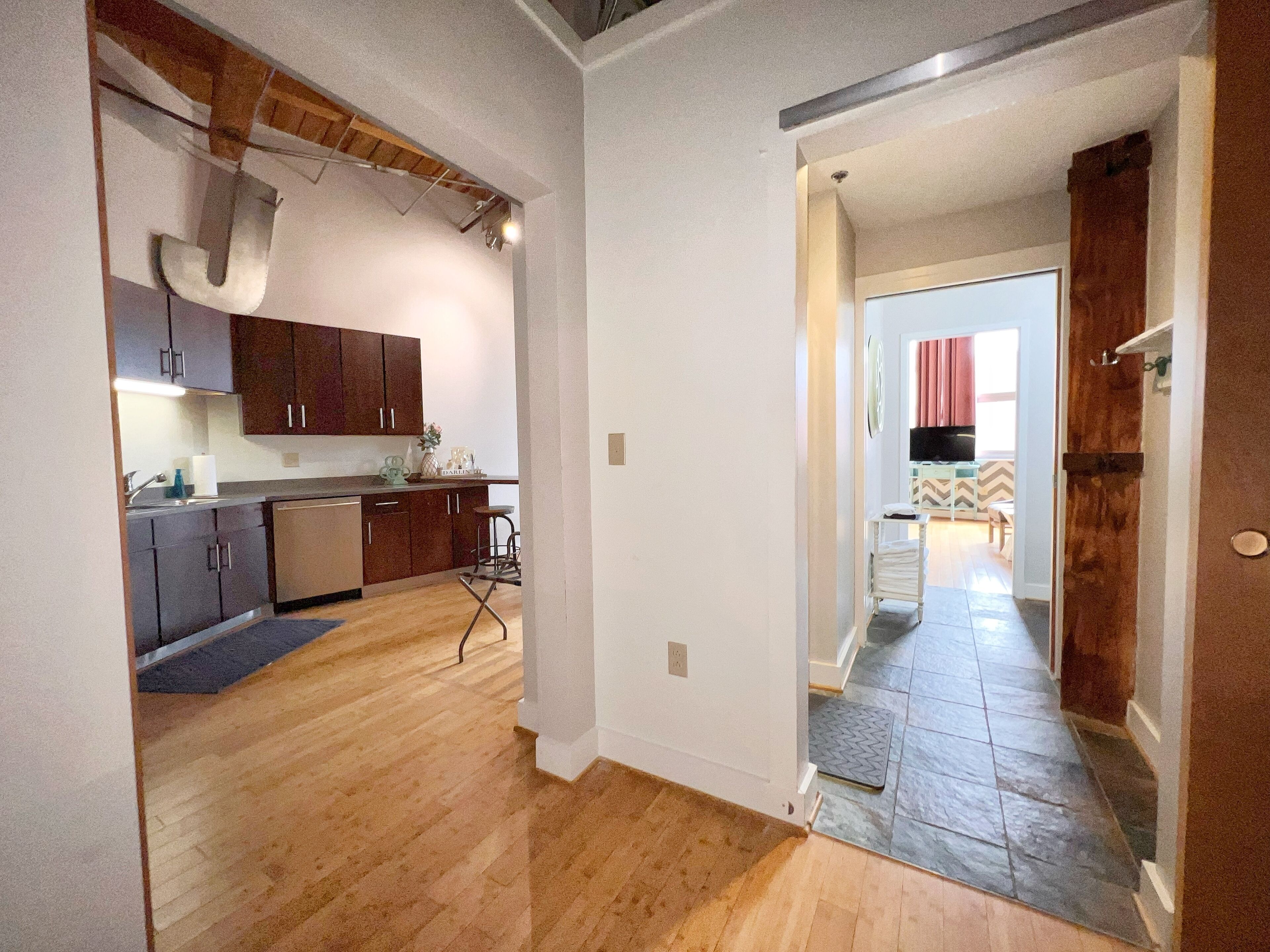 A modern kitchen with wooden cabinets, a dishwasher, and a bar stool. There is a hallway leading to a dining area with a red curtain.