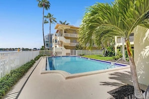 A swimming pool with a white fence, palm trees, and a building with balconies.