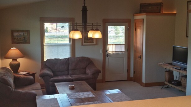 A living room with a brown leather couch, a wooden coffee table, a lamp, a television on a stand, and a door with a view of a parking lot. - Award Winning Cow Creek Suite: Edge-Of-Town Quiet & Convenience With Great Views (Ridgway)