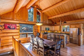 Interior de una cabaña de madera con una gran mesa de comedor, sillas de madera, una zona de cocina y una chimenea.