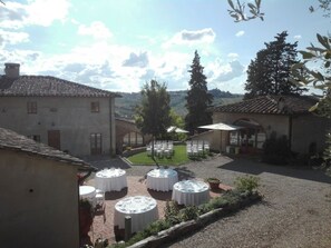 A courtyard with round tables set up for dining, surrounded by buildings with tiled roofs and a grassy area.