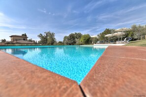 A clear blue swimming pool with wooden deck edges, surrounded by trees and a building with a tiled roof.