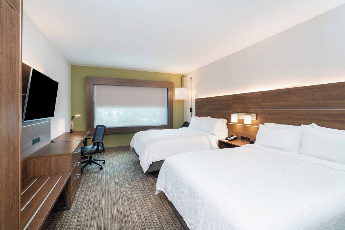 Ein Hotelzimmer mit zwei Betten, einem Schreibtisch mit Stuhl, einem Flachbildfernseher, einem großen Fenster und einer Wandlampe.