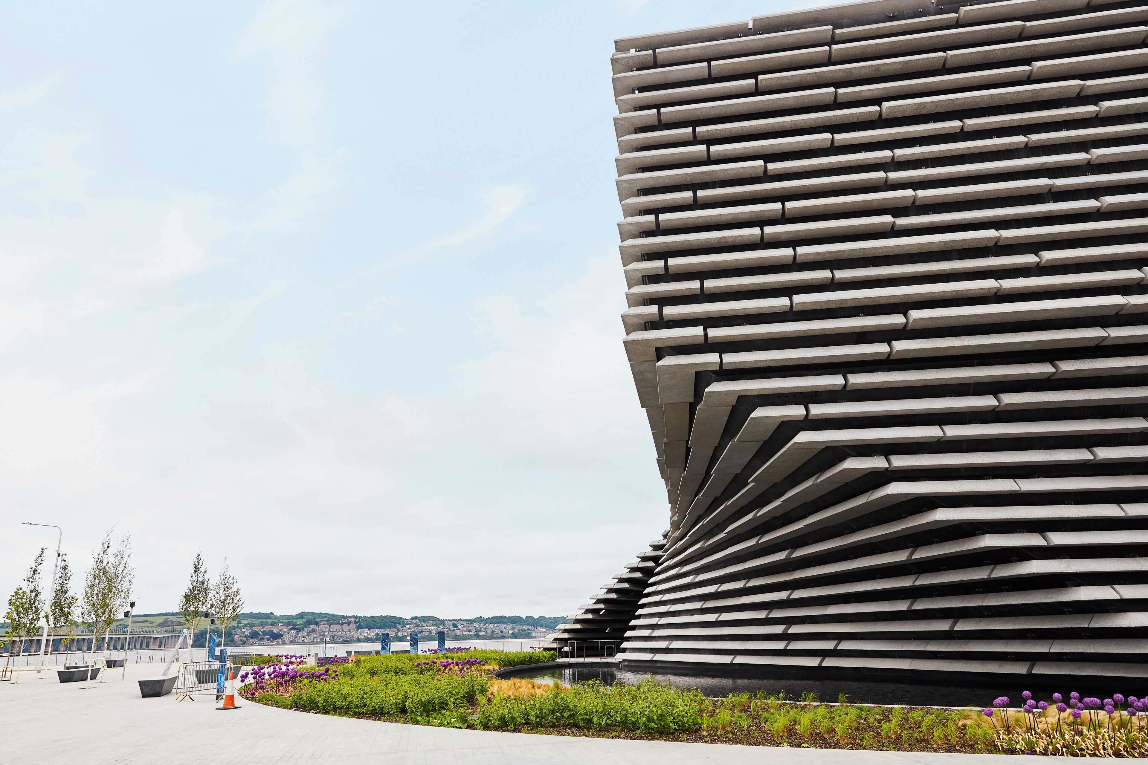 A modern architectural structure with a curved design, surrounded by a landscaped area with greenery and purple flowers.