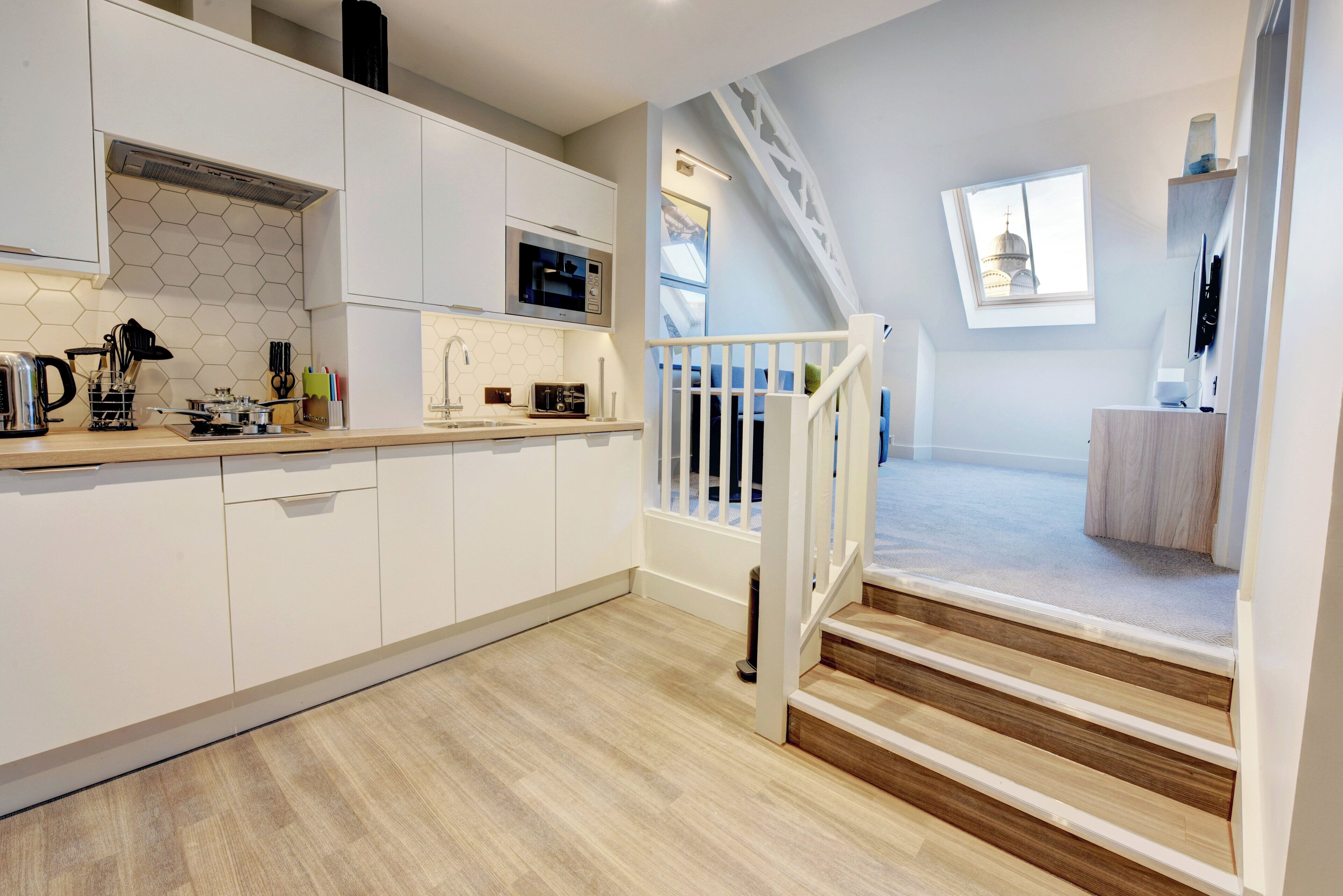 Une cuisine moderne avec des meubles blancs, un four à micro-ondes et un grille-pain. Un escalier mène à un balcon d’où l’on peut admirer un immeuble.
