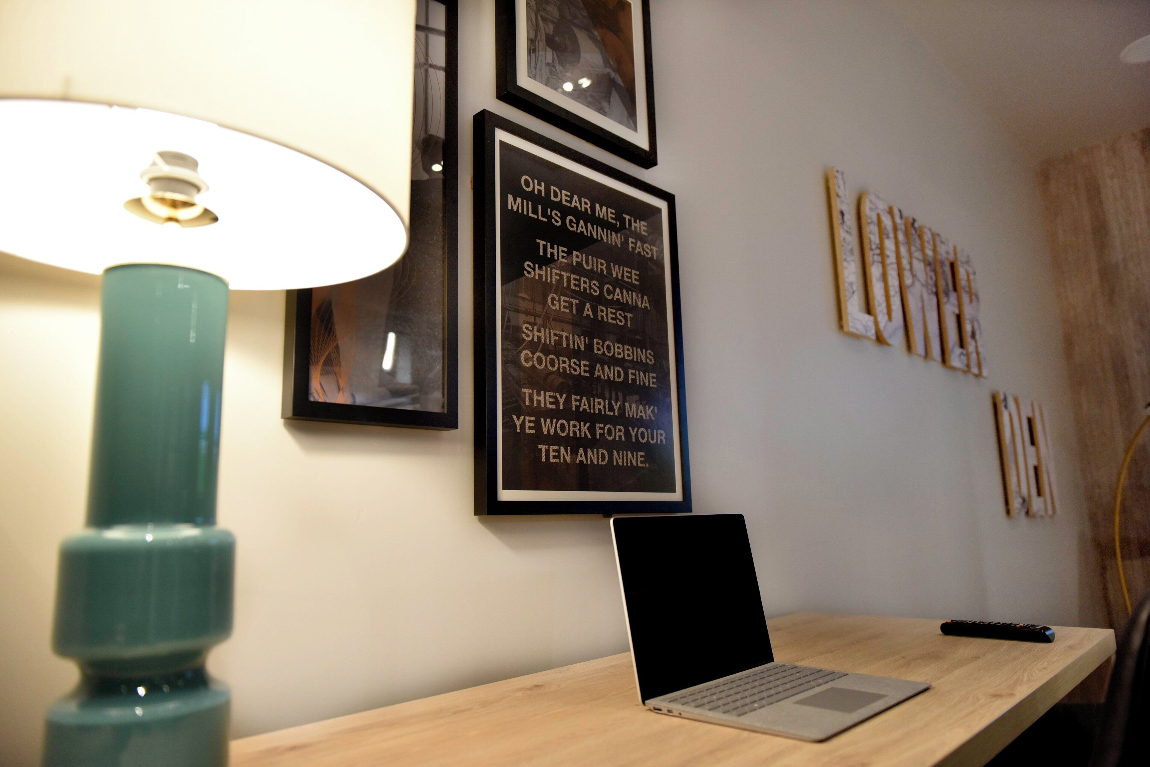 Ein Schreibtisch mit einem Laptop, einer Lampe und gerahmten Text an der Wand.