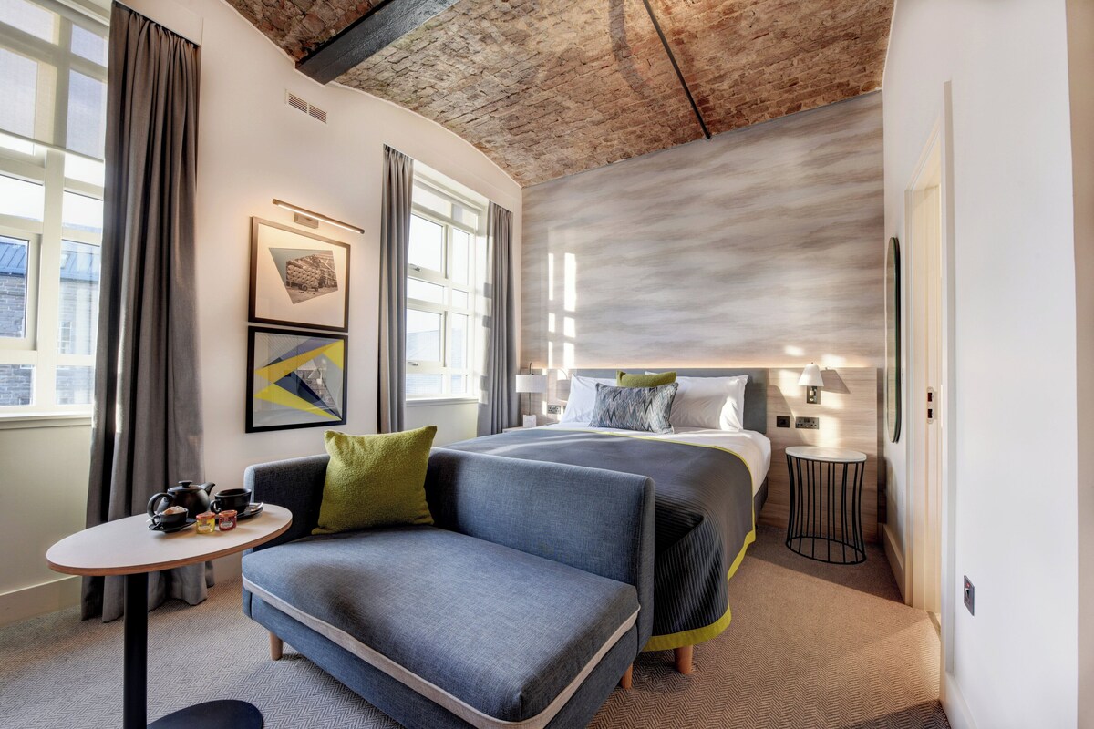 A modern hotel room with a large bed, a sofa, a small table with tea set, and artwork on the walls.
