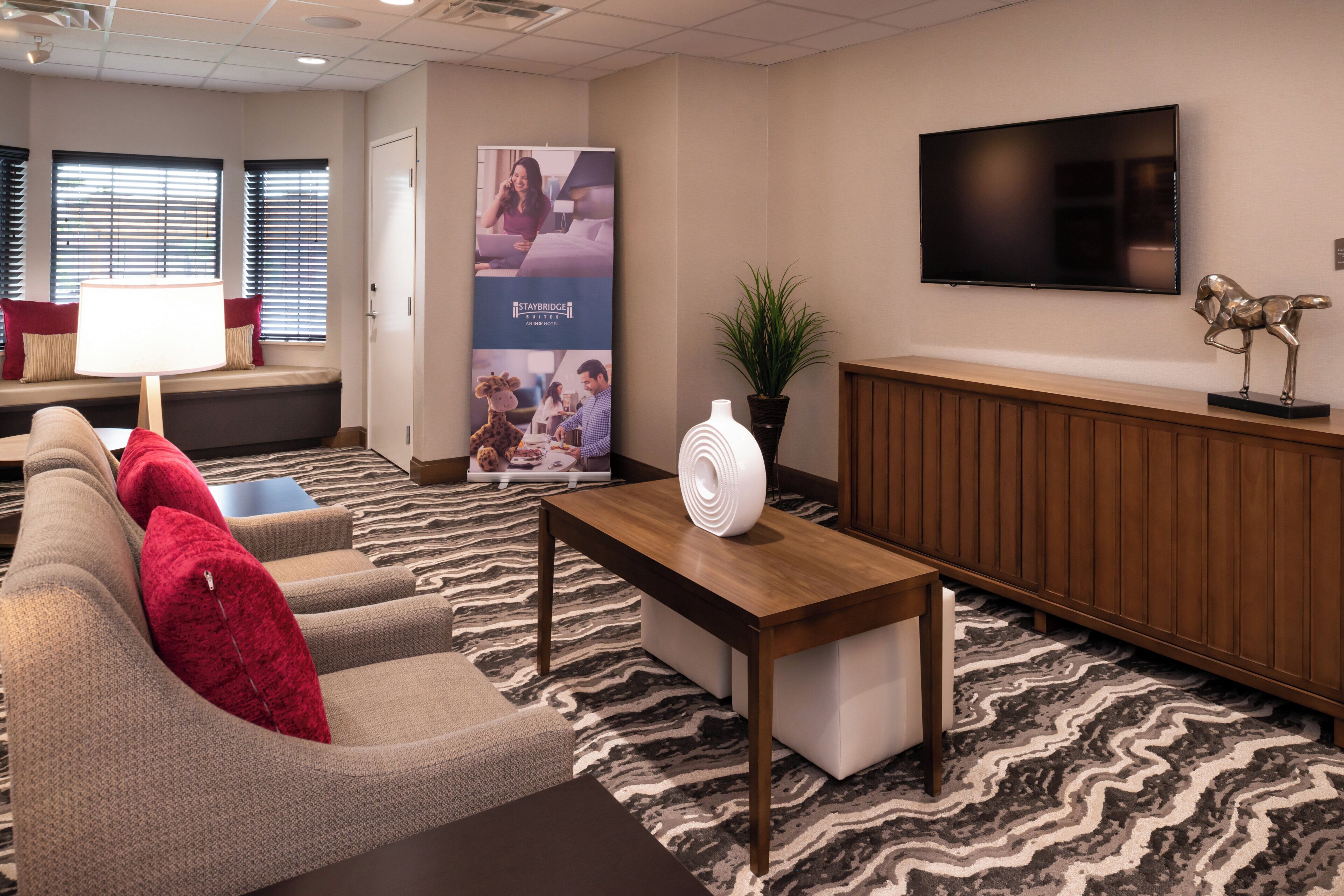 A hotel lobby with a sofa set, a coffee table, a television mounted on the wall, a plant, and a poster.