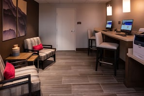 A hotel lobby with a reception desk, two computer monitors, a printer, and a seating area with chairs and a small table.