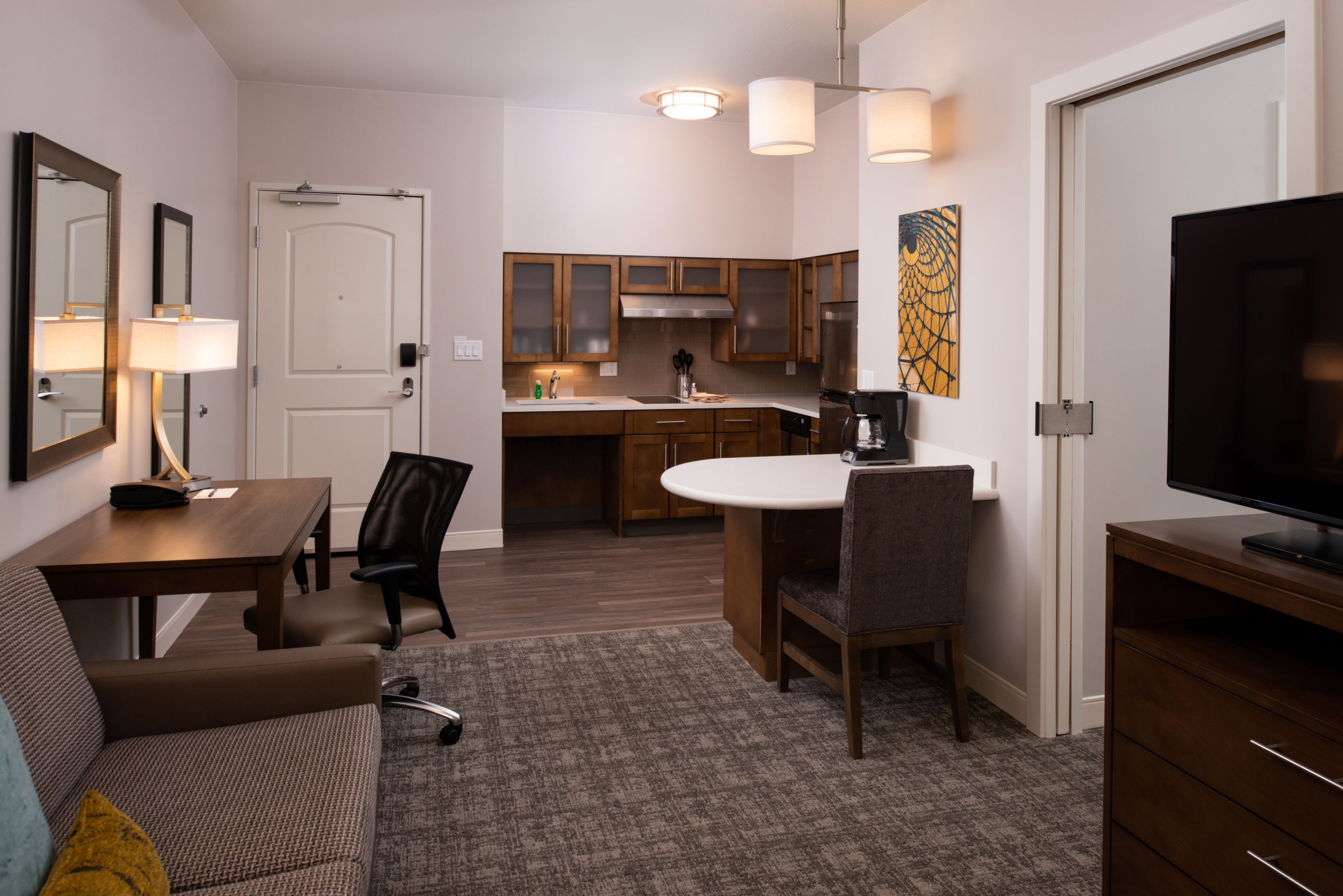 A compact hotel room with a kitchenette, a dining area, a sofa, and a TV.