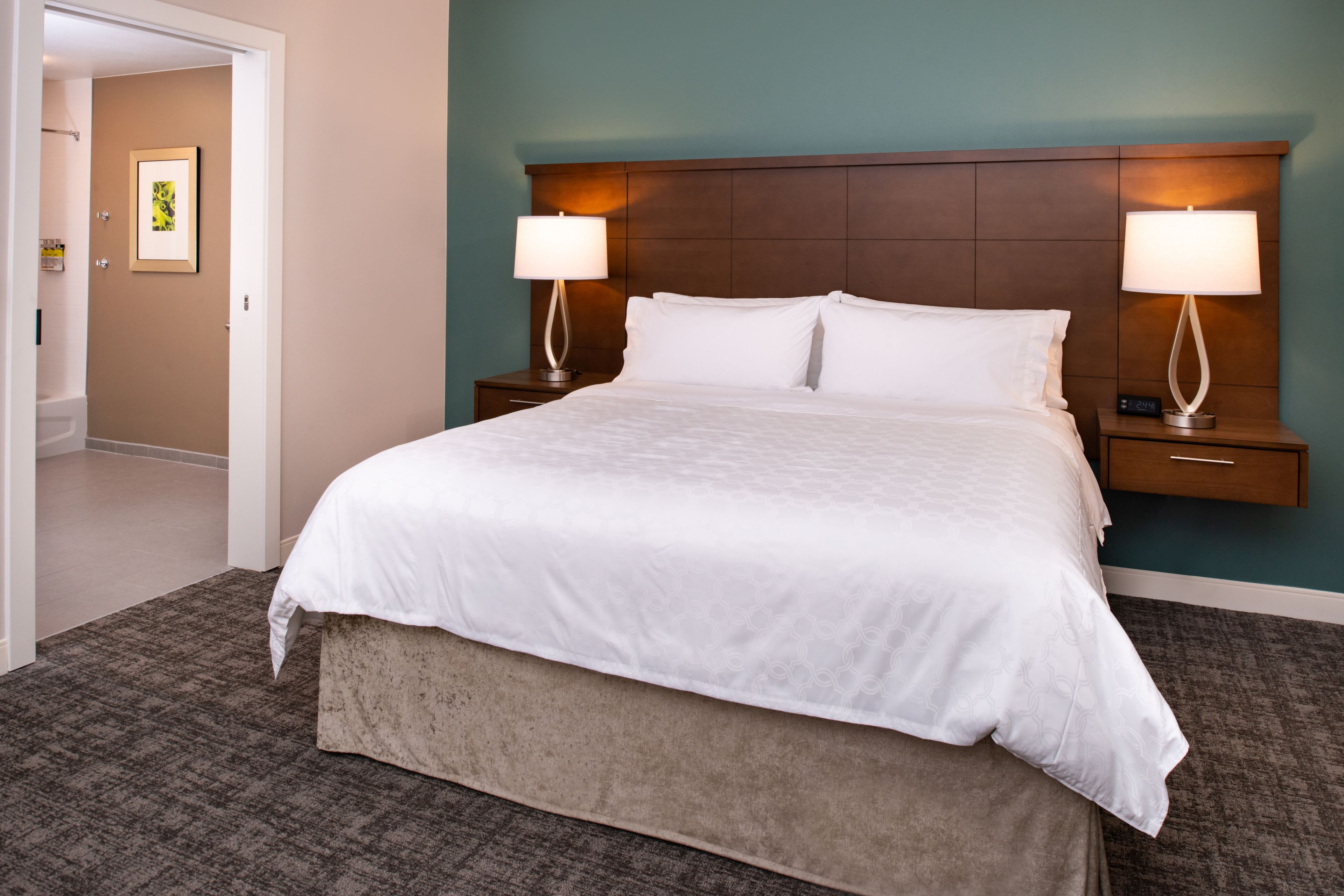 A hotel room with a large bed, two bedside tables with lamps, a wooden headboard, and a carpeted floor.