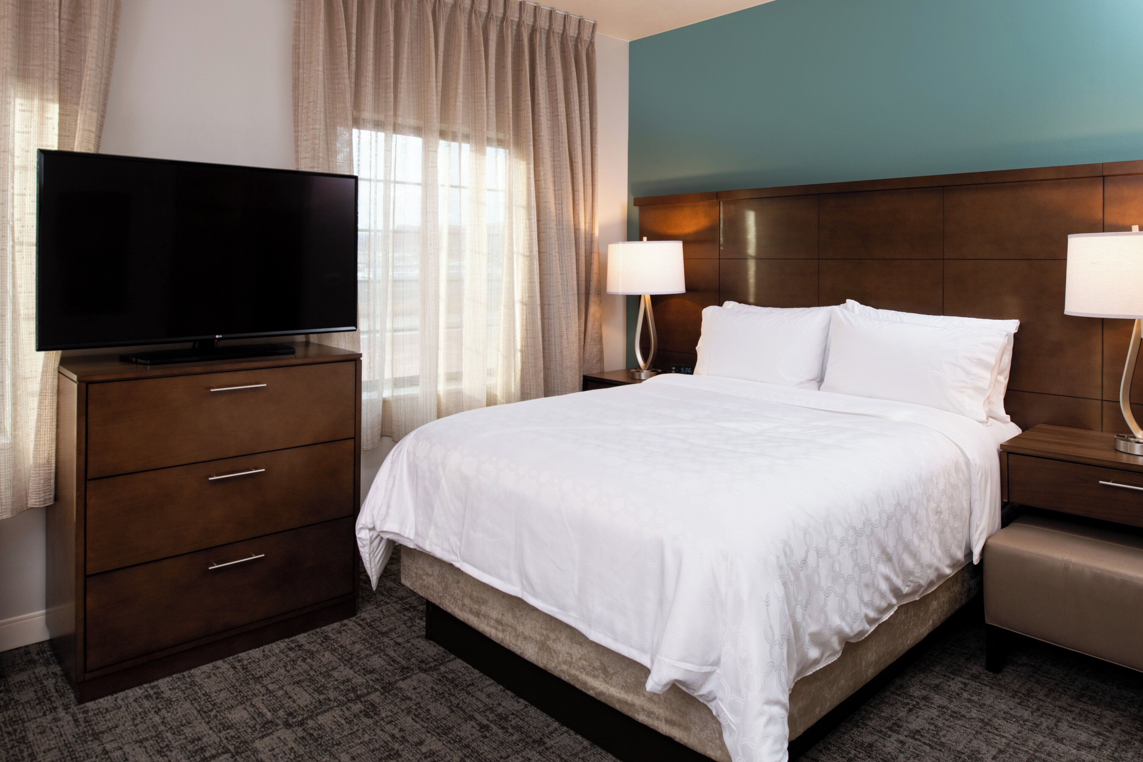A hotel room with a bed, a TV on a dresser, two bedside lamps, and a window with curtains.