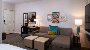 Quarto de hotel com cama, sofá, escrivaninha com cadeira, luminária e obras de arte nas paredes.