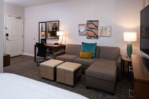 Habitación de hotel con cama, sofá, escritorio con silla, lámpara y obras de arte en las paredes.