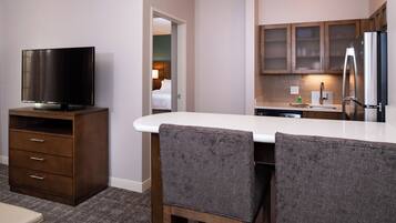 Een hotelkamer met een kitchenette, een flatscreen-tv, een eethoek met twee stoelen, en een bed dat door een open deur zichtbaar is.