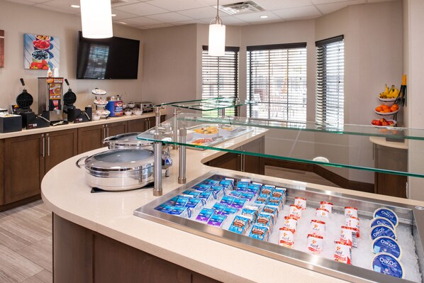 A hotel breakfast area with a self-service station, a television mounted on the wall, and a window with blinds.