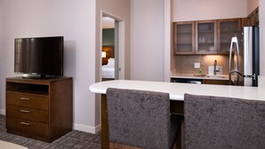 Quarto de hotel com cozinha compacta, televisão sobre um móvel de madeira e duas poltronas em frente a uma mesa branca.