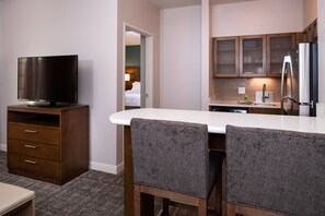 A hotel room with a kitchenette, a TV on a wooden cabinet, and two upholstered chairs at a white table.