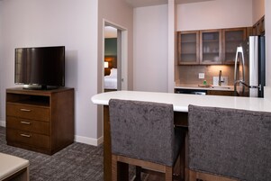 Habitación de hotel con kitchenette, un televisor sobre un mueble de madera y dos sillones tapizados frente a una mesa blanca.